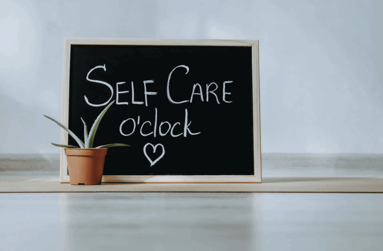 a blackboard with the words "self care o'clock" and a small heart below it, written on it next to a potted plant